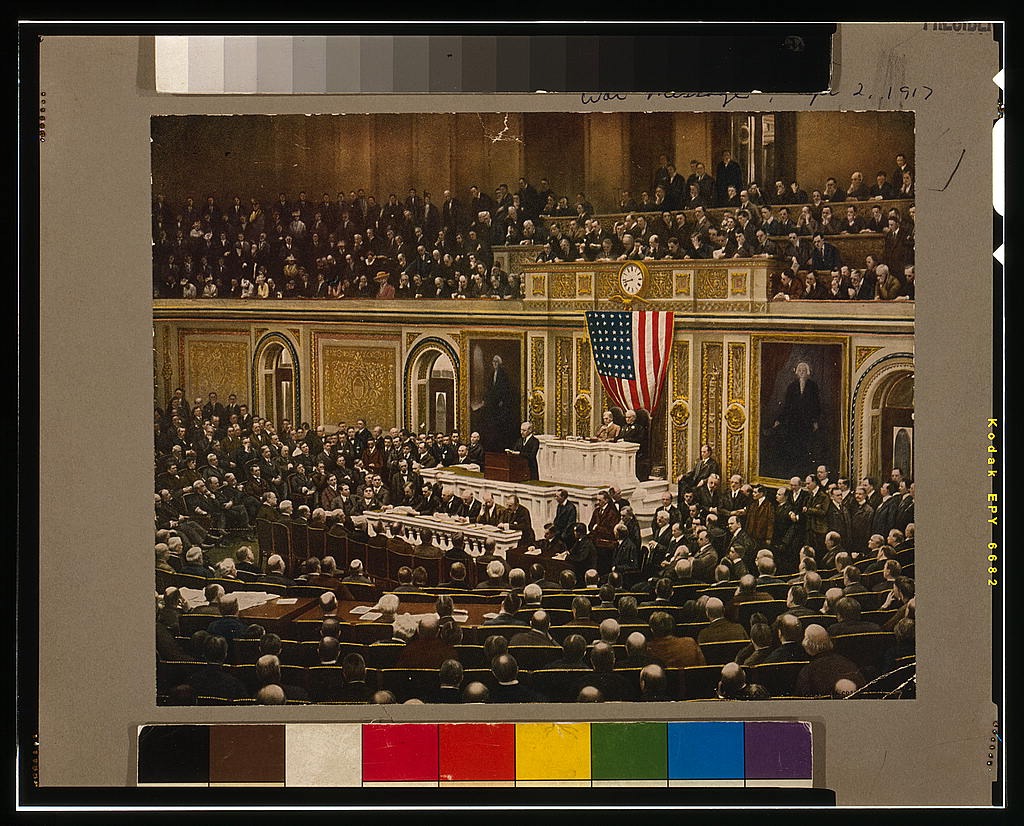 A color halftone print depicting President Woodrow Wilson standing before a joint session of Congress on April 2, 1917, addressing a packed chamber to request a declaration of war against Germany. American flags are visible in the background.