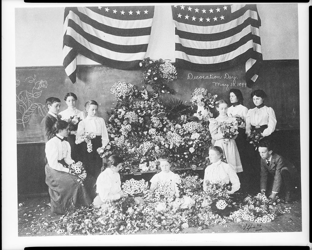 Black-and-white photograph of schoolchildren gathered around a large floral display beneath American flags for a Decoration Day observance on May 30, 1899. Several students hold bouquets and wreaths as part of the remembrance ceremony.