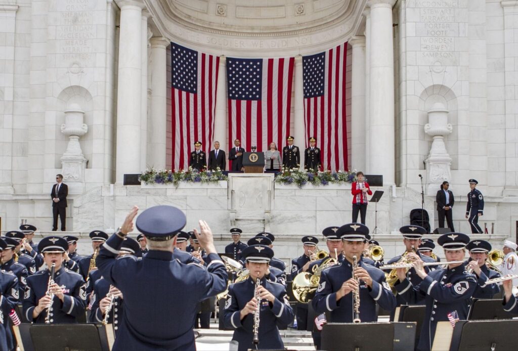 Memorial Day ceremony with military personnel, veterans, and civilians gathered outdoors near wreaths and American flags during a public remembrance event.