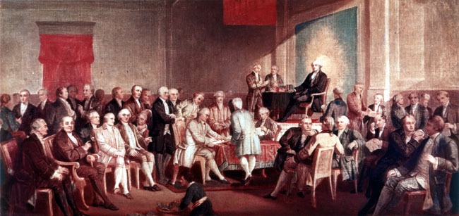 Historical painting showing delegates signing the United States Constitution inside Independence Hall, with parchment documents spread across a table and delegates gathered around.