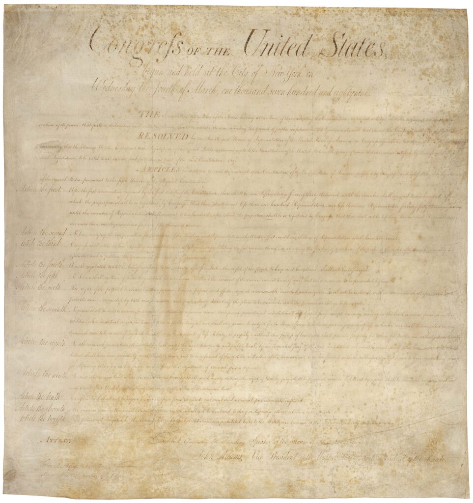 Section of the handwritten Bill of Rights on parchment showing the text of the First Amendment in eighteenth-century script. The document's ink writing appears on aged parchment with visible folds and historical wear.