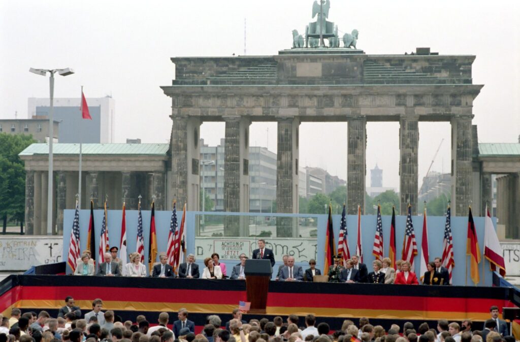 President Ronald Reagan stands at a podium delivering a speech before a large crowd at the Brandenburg Gate in West Berlin. American and West German flags line the stage, while the Berlin Wall and the iconic Brandenburg Gate rise behind him.