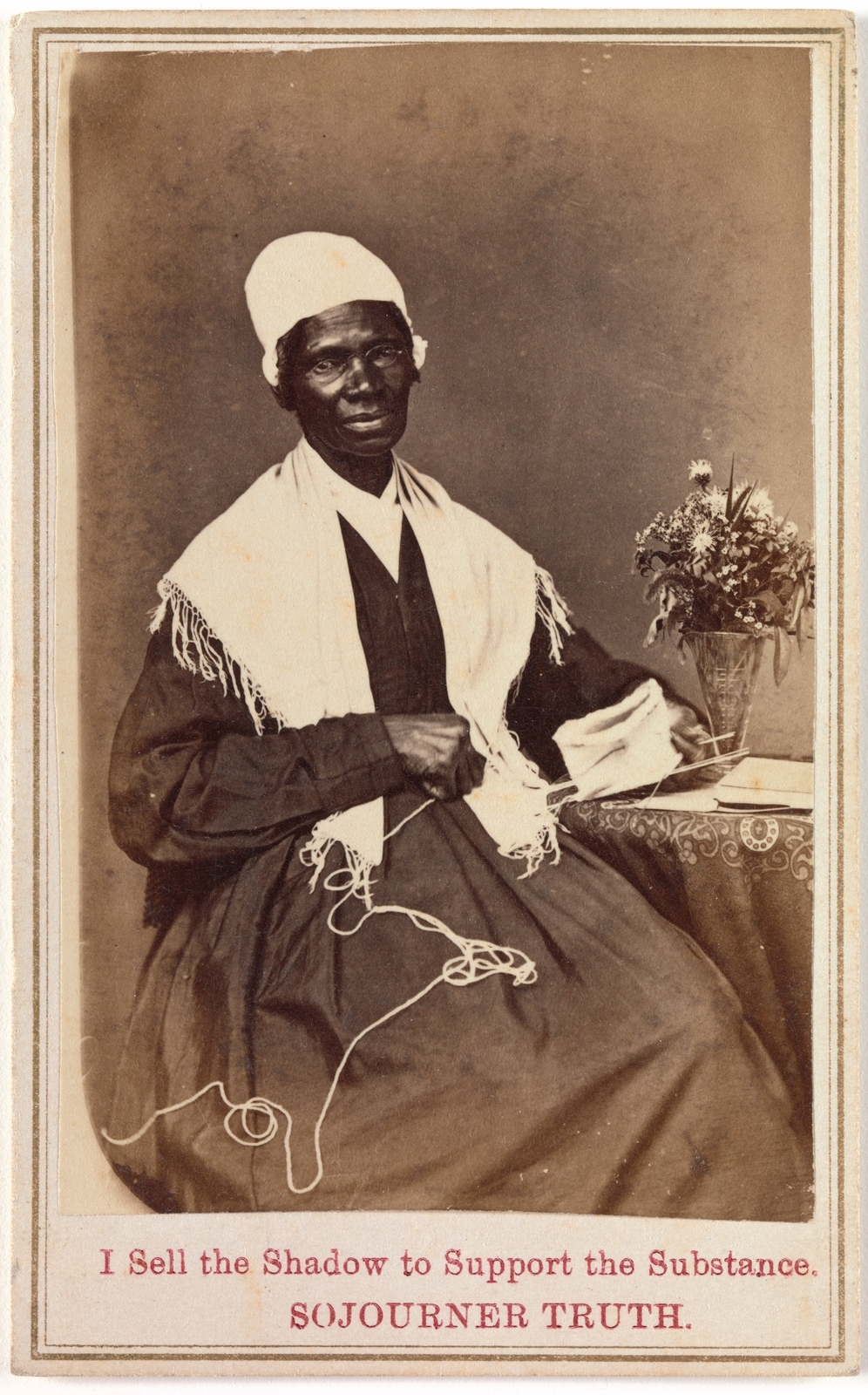 A black-and-white 1864 photograph of Sojourner Truth seated with knitting in her hands, wearing a white head covering and patterned dress.