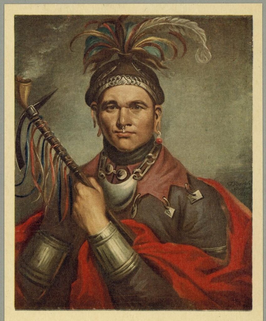 A color half-length portrait of Cornplanter against a gray-green background. He wears a cap crowned with teal, brown, gold, and white feathers and a silver band, a nose ring, long earrings, and a silver chain necklace with a crescent-shaped gorget. A red blanket is draped over a gray-brown coat fastened with silver brooches, and wide silver bands circle his wrist and forearm. He holds a ribboned pipe tomahawk upright, with smoke rising from its bowl.