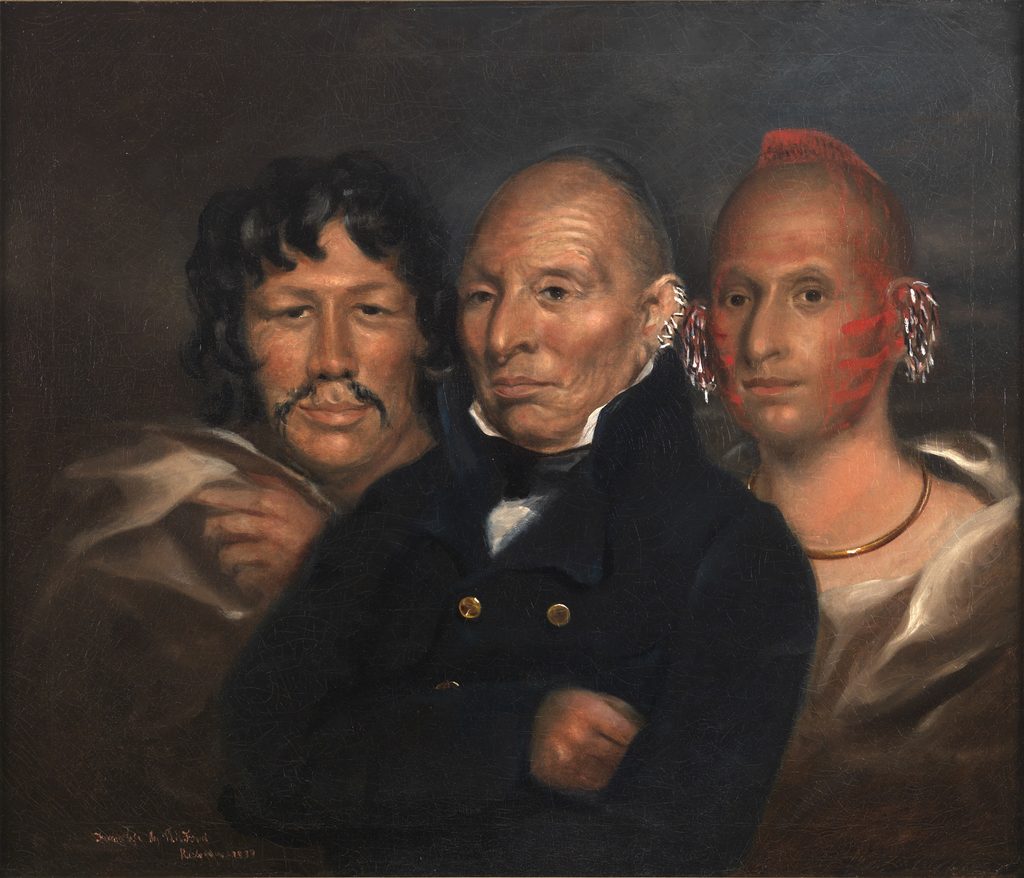 An 1833 oil portrait of three Sauk men against a dark background. At center, Black Hawk, an older man with a shaved head and scalp lock, wearing ear ornaments and a dark double-breasted coat with brass buttons, stands with his arms folded. At left, a man with long dark wavy hair and a mustache holds a light blanket at his shoulder. At right, a younger man with a red roach, red face paint, ear ornaments, and a gold neck ring wears a light blanket. The artist’s inscription and the date 1833 appear at lower left.